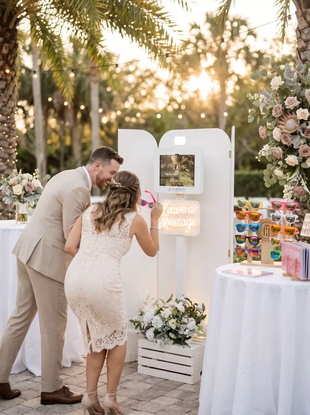 Couple using a photo booth with white panels and neon "Leave a Message" sign at an outdoor event with palm trees, string lights, and floral decorations