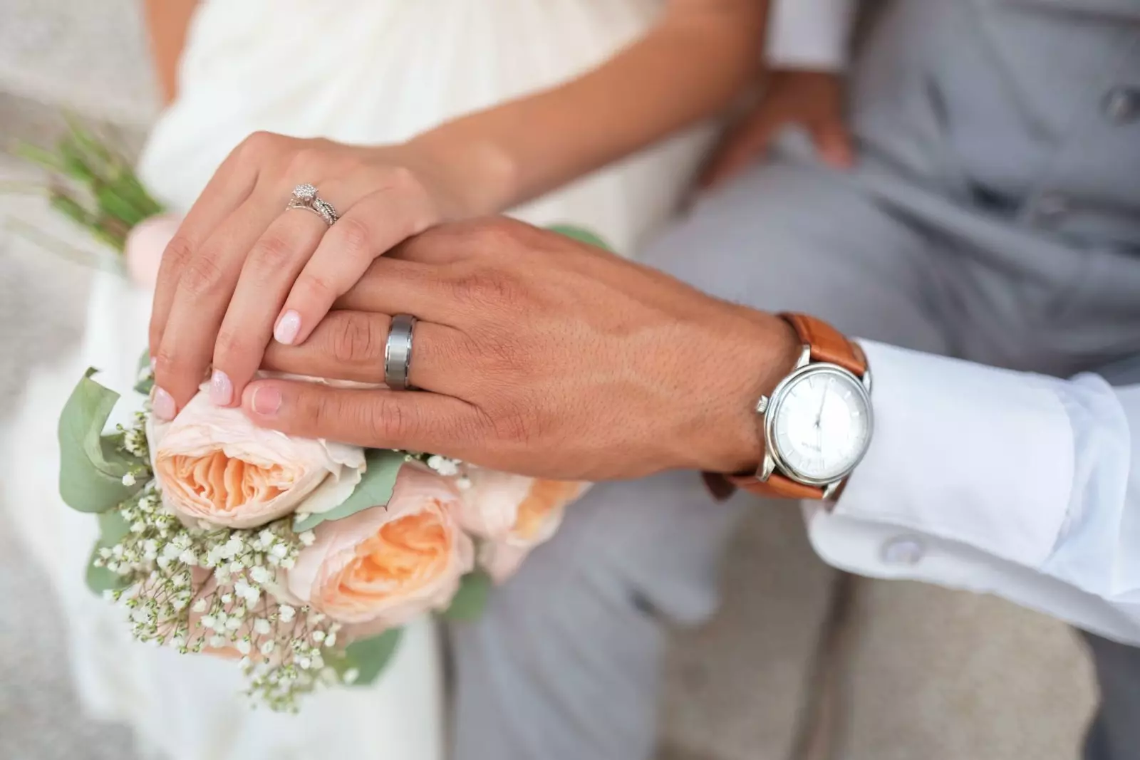 Couple displays their wedding rings while holding a bridal bouquet of peach roses and white flowers as they prepare for wedding planning help