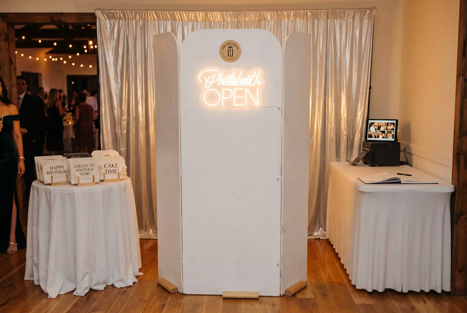 White photo booth with neon "Photobooth OPEN" sign flanked by draped curtains, with guest tables and signage on either side in an event space