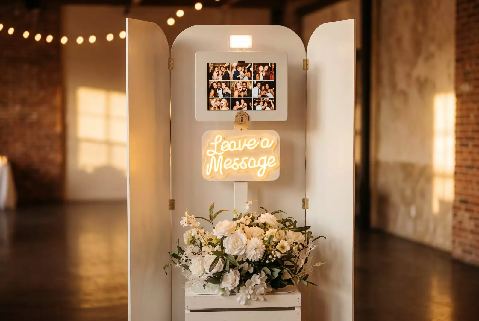 Photo booth with tri-fold enclosure behind it displaying a screen with guest photos above a neon "Leave a Message" sign and floral arrangement below