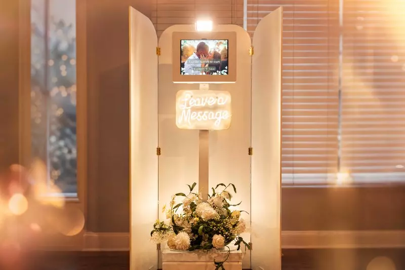 Video Guestbook Photo booth with illuminated neon sign reading "Leave a Message," display screen, and floral arrangement at base, positioned near windows with warm lighting