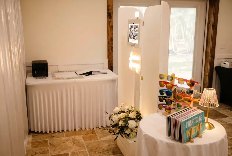 Photo booth setup with white curtained enclosure, attendant table, flower arrangement, and colorful props display in an event venue