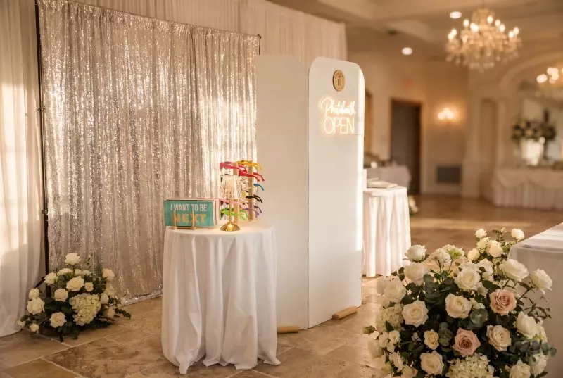 Photo booth setup in an elegant ballroom with white cylindrical booth, sequined backdrop, white-draped table, floral arrangements, and neon open sign